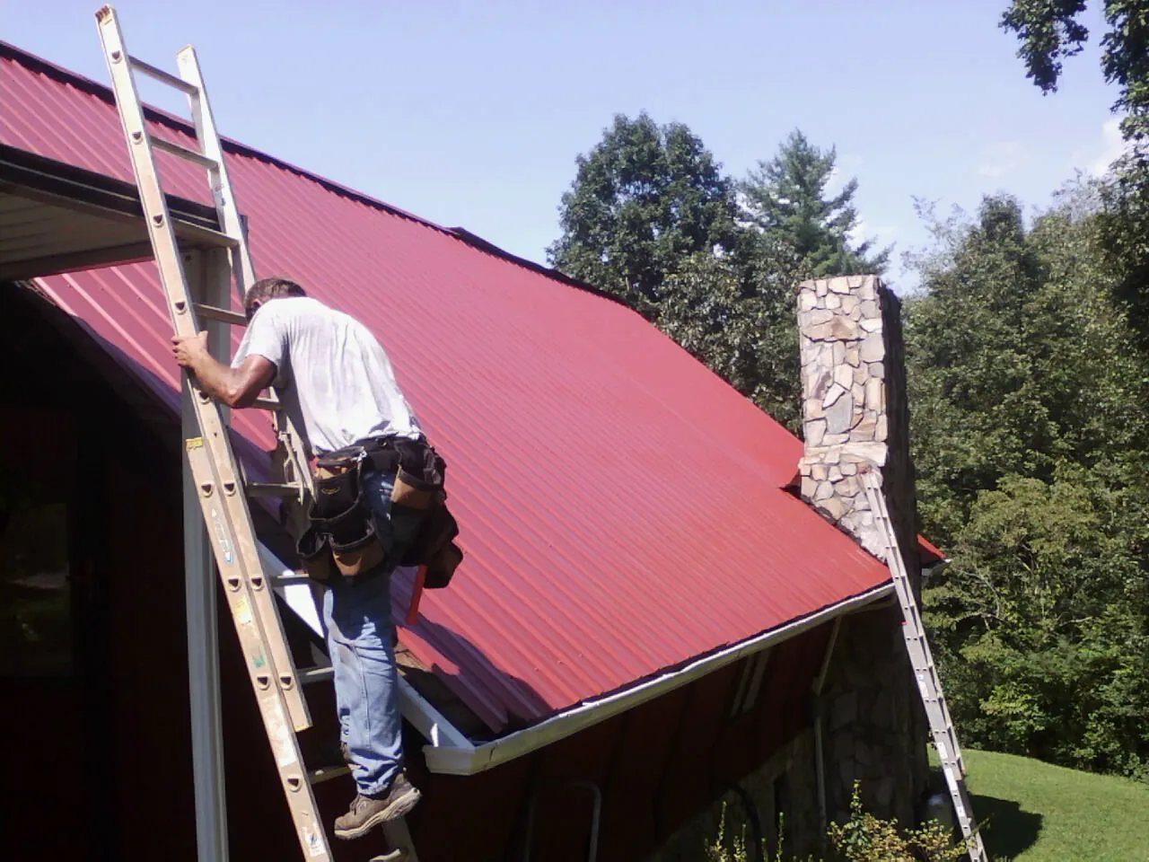 Roofer on a ladder beside a red metal roof and stone chimney — Soffit & Fascia Repair by Roofing Brothers in Wallington